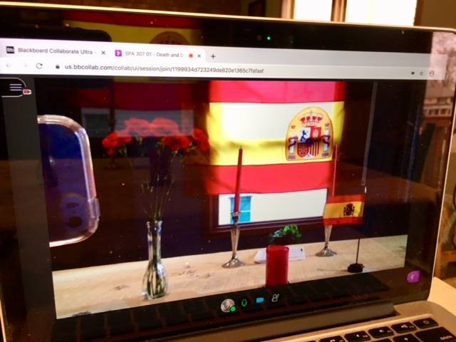 Sigma Delta Pi flag hung up in background with red candles and a vase of red carnations on a table in the foreground as viewed on a computer screen